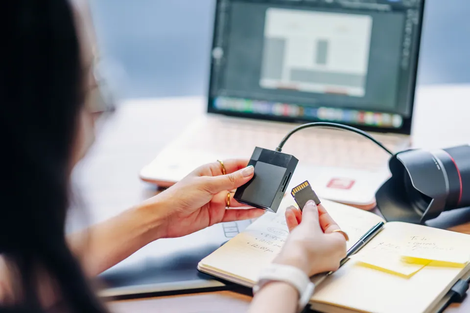 Photographer holding a memory card beside a card reader and laptop